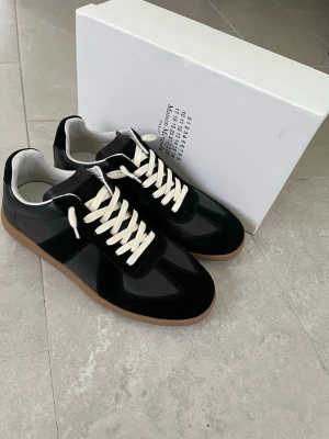 Svarta Maison Margiela Replica sneakers - Snygga svarta Maison Margiela Replica sneakers med vita skosnören och gummisula. Ovandelen är i en mix av mocka och läder, med klassisk låg silhuett och diskret logga på plösen. Perfekta för dig som gillar stilrena och tidlösa sneakers.