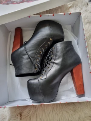 Jeffrey Campbell Lita - Säljer ett par svarta stövletter med snörning och kraftig platåsula. Skorna har höga, breda klackar i träfärg och guldfärgade öljetter. Tillverkade i konstläder med en edgy och trendig look. Perfekta för dig som vill sticka ut och älskar statementskor.