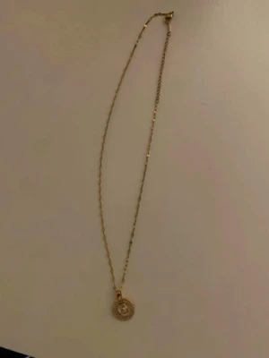 Guldigt halsband med cirkelberlock - Säljer ett elegant halsband i guldfärg med en tunn kedja och en rund berlock. Berlocken har en cirkelformad design med små detaljer runt kanten och en klar sten i mitten. Perfekt för dig som gillar stilrena smycken med en twist.