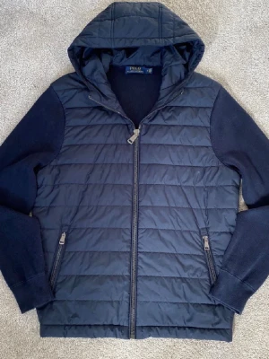 Marinblå cardigan Polo Ralph Lauren - Snygg marinblå jacka från Polo Ralph Lauren med quiltad framsida, stickade ärmar och huva. Jackan har dragkedja framtill och två fickor med dragkedja. Perfekt för kyliga dagar och enkel att matcha med olika outfits. Nästan oanvänd säljer pga för många jackor i garderoben.