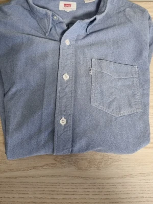 Blå skjorta från Levi's i bomull - Klassisk blå skjorta från Levi's i mjuk bomull. Skjortan har knappar framtill, en bröstficka och en enkel, stilren design. Perfekt till jeans eller chinos för en avslappnad look.