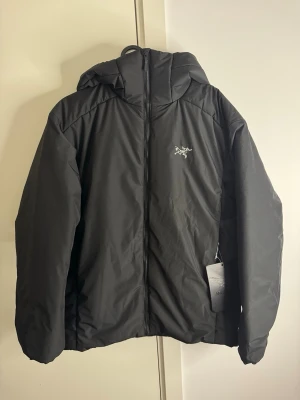 Svart pufferjacka från Arc'teryx ATOM HOODY - Svart pufferjacka från Arc'teryx med huva och dragkedja framtill. Jackan har ett stilrent broderat logotyptryck på bröstet och är tillverkad i ett slitstarkt material som håller värmen under kalla dagar. Perfekt för dig som vill ha en snygg och funktionell vinterjacka.