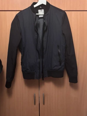 Mörkblå  J.Lindberg cardigan - Snygg mörkblå cardigan. Jackan har två fickor med dragkedja och ett coolt mönstrat innerfoder. Perfekt för de uppkommande vårdagarna. 