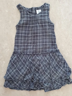 Klänning - Säljer en grå ärmlös klänning från H&M med vitt rutmönster. Klänningen har volang nedtill och är tillverkad i mjuk bomull, perfekt för en avslappnad och trendig look.