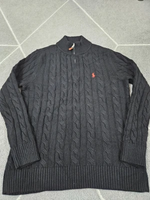 Svart kabelstickad tröja Polo Ralph Lauren - Kabelstickad svart tröja från Polo Ralph Lauren med halv dragkedja och röd broderad logga på bröstet. Tröjan har ribbade muddar och hög krage. Perfekt för dig som gillar klassisk och stilren design.