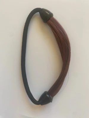 Hårsnodd med rödbrunt syntethår - Elastisk hårsnodd med fastsatt rödbrunt syntethår. Perfekt för att snabbt fixa en snygg hästsvans eller fläta med extra volym och färg. Hårsnodden är svart och har plastfästen i ändarna. Passar dig som vill ha en enkel men effektfull håraccessoar.