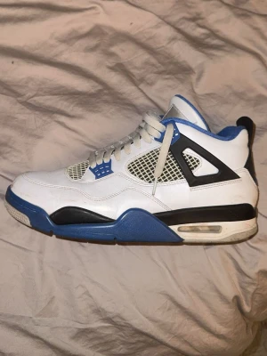 Air Jordan 4 Retro White/Blue - Säljer ett par Air Jordan 4 Retro sneakers i vitt läder med blå och svarta detaljer. Skorna har meshpaneler på sidorna, synlig Air-enhet i sulan och klassisk Jumpman-logga på hälen. Feta snören och ikoniska plastdetaljer vid snörningen ger en riktigt clean look.