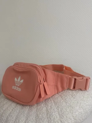 Rosa magväska från Adidas - Snygg magväska från Adidas i en fräsch ljusrosa färg. Väskan har två dragkedjor, justerbart midjeband och klassisk Adidas-logga framtill. Perfekt för att bära det viktigaste och hålla händerna fria. Tillverkad i slitstark textil.