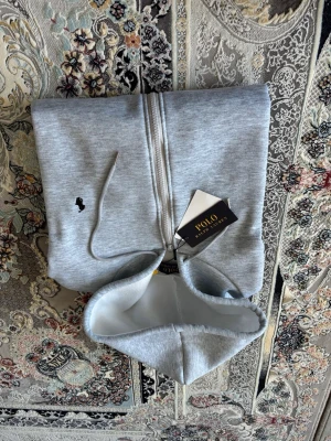 Ljusgrå hoodie från Polo Ralph Lauren - Snygg ljusgrå hoodie från Polo Ralph Lauren med dragkedja och klassisk huva. Broderad svart logga på bröstet och justerbara snören i huvan. Mjuk bomullsblandning som känns skön mot huden. Perfekt för en avslappnad och stilren look.