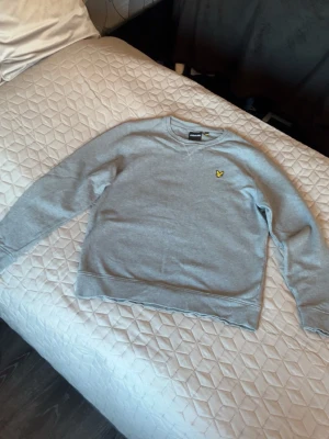 Grå sweatshirt från Lyle & Scott - Snygg grå sweatshirt från Lyle & Scott med klassisk rund hals och gul logga på bröstet. Tröjan har ribbade muddar vid ärmslut och nederkant, perfekt för en avslappnad och stilren look. L / M. Aldrig använd