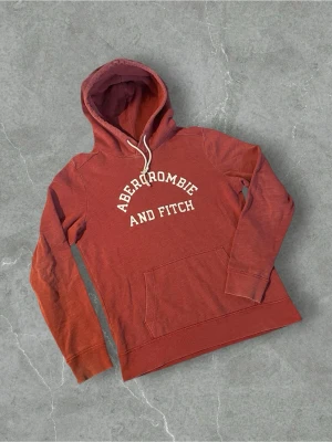 Röd hoodie från Abercrombie & Fitch | S - Snygg röd hoodie från Abercrombie & Fitch med vit logga tryckt på bröstet. Hoodien har en klassisk känguruficka och vit dragsko i huvan. Perfekt för en avslappnad och trendig stil. Mjuk bomullsblandning som känns skön mot huden.
