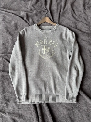 Grå tröja med tryck från Morris - Snygg grå tröja från Morris med rund halsringning och ribbade muddar. Framsidan har ett stort tryck med logga och texten 'Morris' samt ett emblem. Perfekt för en avslappnad och stilren look.