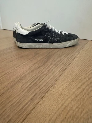 Marinblå Premiata sneakers i mocka - Stilrena svarta sneakers från Premiata med vita detaljer och snörning. Ovandelen är i mocka och läder, med tydlig logga på sidan och hälen. Sulan är vit med diskret mönster. Perfekta för dig som gillar en clean och klassisk look.