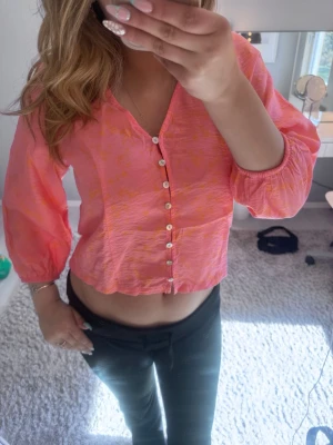 Rosa blus med knappar och ballongärm - Trendig rosa blus med orange mönster och pärlemorknappar framtill. Blusen har v-ringning och trekvartsärm med resår vid ärmslut. Materialet är tunt och lätt, perfekt för varmare dagar. Snygg att styla till både jeans och kjol.