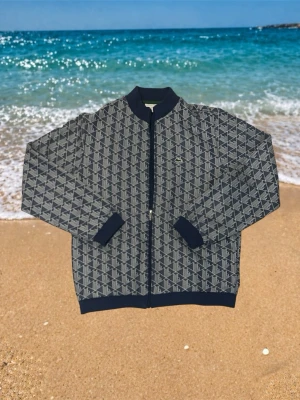  Lacoste tröja - Snygg bomberjacka från Lacoste med mörkblå muddar och hel dragkedja. Jackan har ett grått och blått geometriskt mönster samt den klassiska krokodilloggan på bröstet. Perfekt för dig som vill sticka ut med stilren streetwear.
