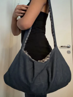 Mörkblå axelväska i tyg - Säljer en mörkblå axelväska i mjukt tyg med bred axelrem. Väskan har en rundad form och insidan är fodrad med ett rutigt tyg i blått och vitt. Perfekt för dig som vill ha en enkel och rymlig väska med coolt mönstrat foder.