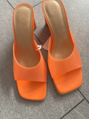 Orange wedgesandaler från H&M - Snygga orange wedgesandaler från H&M med bred rem och fyrkantig tå. Klacken är hög och kilformad, vilket ger både höjd och stabilitet. Materialet är glansigt och plastigt, vilket ger en modern vibe. Perfekta för dig som vill sticka ut med färg.
