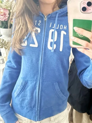 Blå zip-hoodie Hollister - Blå hoodie med dragkedja från Hollister, med vita tryckta siffror och text på bröstet. Utrustad med huva och dragsnören samt två fickor framtill. Mjuk bomullsmix och ribbade muddar vid ärmslut och nederkant. 