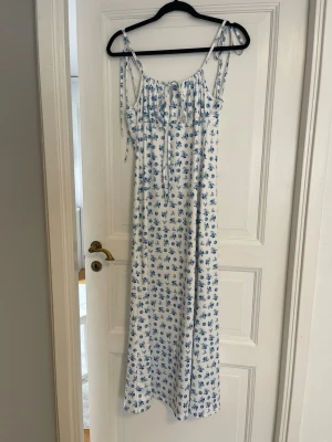 Vit långklänning med blå blommor - Superfin långklänning med smala axelband och knytning upptill. Klänningen är vit med blått blommigt mönster och har en luftig, rak passform. Perfekt för sommarens varma dagar och har en romantisk vibe. Materialet känns lätt och bekvämt. Klänning anpassar sig till din kropp och axelbanden är anpassningsbara.