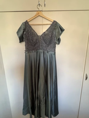 Grå långklänning med spetsdetaljer - Elegant grå långklänning med v-ringad spetsöverdel och korta ärmar. Klänningen har vackra broderade detaljer och paljetter på överdelen, samt ett flödande satinliknande kjolparti. Perfekt för dig som vill sticka ut med en lyxig look. Passar storlekar M-XL då den har snörning. Nypris 2000kr.