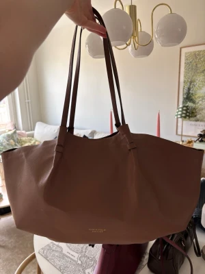 Brun shoppingväska - Stilren shoppingväska i brun färg från H&M. Väskan har en rymlig och mjuk form med dubbla långa axelband och är tillverkad i slätt läder. Perfekt för dig som vill ha en enkel och snygg väska med plats för allt du behöver.
