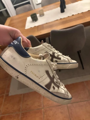 Feta Premiata Skor - Snygga vita sneakers från Premiata med blå detaljer och märkets logga på hälen. Skorna har lågt skaft och är tillverkade i läder med gummisula. Klassisk design med subtila grå inslag och tryck på sulan. Perfekta för en stilren och trendig look. Ny pris 3500kr mitt pris 799💸🍾