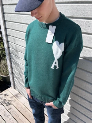 Grön stickad tröja från Ami Paris - Snygg grön stickad tröja från Ami Paris med stort vitt hjärta och bokstaven A på bröstet. Tröjan har rund halsringning och ribbade muddar. Perfekt för dig som gillar stilrena och trendiga plagg med en twist.