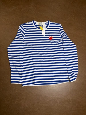 Randig långärmad topp Comme des Garçons - Blåvit randig långärmad topp från Comme des Garçons Play med klassisk rund hals och rött hjärta broderat på bröstet. Tillverkad i mjuk bomull och perfekt för en stilren och trendig look.