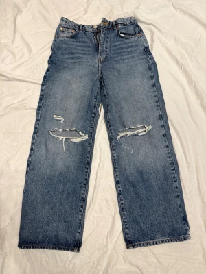 Blå raka jeans med slitningar - Säljer ett par blå jeans med raka ben och tydliga slitningar över knäna. Jeansen har klassisk femficksdesign och hög midja. Perfekta för en avslappnad och trendig streetstil.