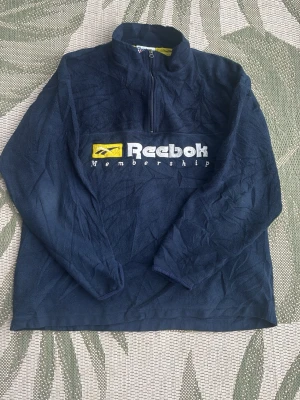 Mörkblå fleece från Reebok - Mysig mörkblå fleece från Reebok med halv dragkedja och broderad logga samt texten 'Membership' på bröstet. Tröjan har hög krage och är tillverkad i mjukt fleecematerial. Perfekt för kalla dagar och sportig stil. Står storlek XL men är liten i storlek så passar storlek L
