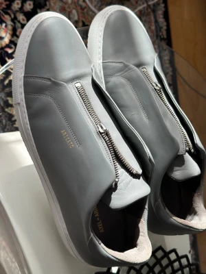 Grå sneakers från Axel Arigato - Stilrena grå sneakers från Axel Arigato i slätt läder med silverfärgad dragkedja på ovansidan istället för klassiska snören. Skorna har vit sula och minimalistisk design med diskret logga i guld på sidan.