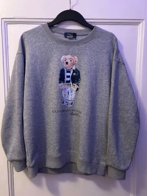 Grå sweatshirt Polo Bear Ralph Lauren - Grå sweatshirt från Polo Ralph Lauren med ikoniskt Polo Bear-tryck på bröstet. Rund halsringning, ribbade muddar och avslappnad passform. Perfekt för en chill och stilren look. Materialet är mjukt och bekvämt, troligtvis bomullsblandning. Det enda problemet med tröjan är att tagen inte är kvar (sista bilden) annars är tröjan helt oskadad. Fråga gärna om ni har några funderingar på något.👍🏼 Mvh Linus😇