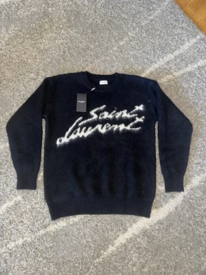 Svart Saint Laurent stickad tröja - Snygg svart stickad tröja från Saint Laurent med vit logotyp i handskriven stil på bröstet. Tröjan har rund halsringning, långa ärmar och är gjord i ett fluffigt, mjukt material som känns lyxigt och trendigt. Det står storlek S men passar till storlek M