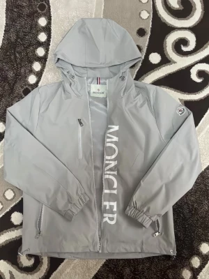 Grå vindjacka från Moncler - Snygg grå vindjacka från Moncler med huva och dragkedja framtill. För fett kvalitet i top notch, det exklusiva jackan passar i every day use.