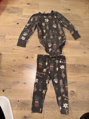 Brunt blommigt bodyset med byxa - Sött set med långärmad body och matchande byxa i brunt tyg med blommor och krukväxter. Bodyn har tryckknappar i grenen och muddar vid ärmslut. Mjukt och stretchigt material, perfekt för en bekväm vardagslook.