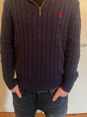 Mörkblå kabelstickad tröja Polo Ralph Lauren - Kabelstickad mörkblå tröja från Polo Ralph Lauren med halv dragkedja och liten röd logga broderad på bröstet. Tröjan har hög krage och är perfekt för lager-på-lager. Klassisk och tidlös design som funkar till det mesta. Kan gå ner i pris på en snabb affär
