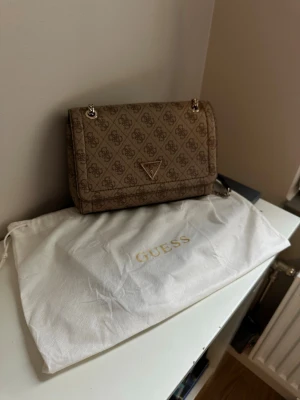 Brun handväska från Guess - Snygg brun handväska från Guess med klassiskt monogrammönster och metallkedja som axelband. Väskan har en triangelformad logga på framsidan och medföljer en vit dustbag med Guess-tryck. Perfekt för dig som gillar trendiga accessoarer.