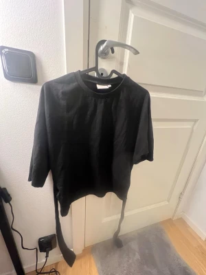 Svart topp med knytband - Cool svart topp med knytband i midjan och rund halsringning. Toppens material känns mjukt och stretchigt, perfekt för att skapa en snygg siluett. Ärmarna är lite längre än vanliga kortärmade och modellen är croppad. Passar dig som gillar stilrena och enkla plagg.