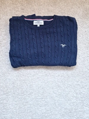 Mörkblå kabelstickad tröja Gant - Mörkblå kabelstickad tröja från HAMPTON REPUBLIC med rund halsringning och broderad logga på bröstet. Tröjan har långa ärmar och klassisk passform. Perfekt för lager-på-lager och enkel att matcha med jeans. Storlek 158/164