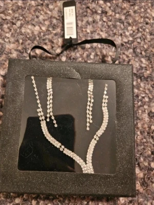 Glittrigt silverset halsband & örhängen - Säljer ett glittrigt smyckeset med matchande halsband och långa örhängen. Setet är täckt av små gnistrande stenar i silverfärg och har en elegant, böjd design. Perfekt för dig som vill sticka ut och addera extra bling till din look.  Kan även hämtas I Partille  