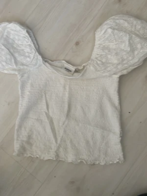 Vit topp med puffärmar från Dobber - Supersöt vit topp från Dobber med stora puffiga kortärmar i broderad spets och smockad kropp. Perfekt för dig som gillar romantisk och trendig stil. Söt volangkant nertill och rund halsringning.