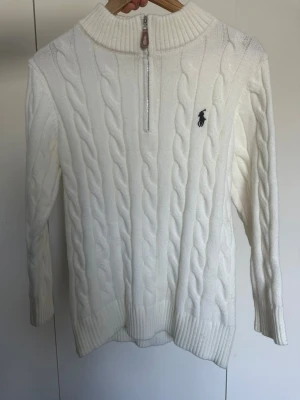 Vit kabelstickad tröja Polo Ralph Lauren - Vit kabelstickad tröja från Polo Ralph Lauren med halv dragkedja och hög krage. Tröjan har lång ärm, ribbstickade muddar och den klassiska broderade loggan på bröstet. Perfekt för lager-på-lager och snygg till jeans.