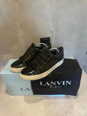 Svarta Lanvin sneakers i mocka och läder - Snygga svarta sneakers från Lanvin i storlek uk5 med ovandel i mocka och detaljer i blankt läder framtill. Klassisk låg modell med vita sulor och svarta skosnören. Perfekta för dig som gillar stilrena och exklusiva sneakers. skorna är i mycket använt skick men har frf en del kvar o ge, klassiskt lanvin problem med skav i hälen, går enkelt o laga. skador och fläckar på låda och fläckar på dustbag men inget märkvärdigt. 