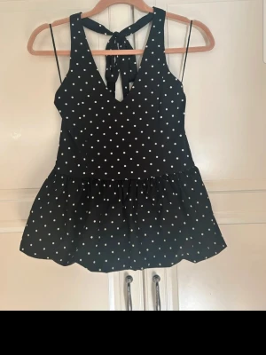 Svart prickig blus med knyt i ryggen - Snygg svart blus med vita prickar och v-ringning. Toppen har tunna axelband och ett knyt i ryggen som ger en extra detalj. Volang nedtill och ärmlös design gör den perfekt för varma dagar. Passar dig som gillar en lekfull och trendig stil.