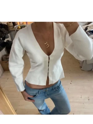 Vit v-ringad kofta med knappar - Snygg vit kofta med djup v-ringning och knäppning framtill. Modellen är kort och har långa ärmar, perfekt att matcha med jeans. Materialet ser ut att vara mjukt och stretchigt, vilket ger en skön passform.