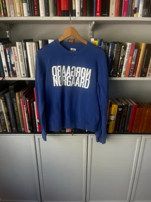 Blå sweatshirt från Mads Nørgaard - Säljer en blå sweatshirt från Mads Nørgaard med vit, spegelvänd logga tryckt på bröstet. Tröjan har rund halsringning, ribbade muddar vid ärmslut och nederkant. Perfekt för en avslappnad och trendig look.
