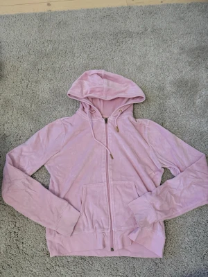 Ljusrosa hoodie Gina tricot - En ljusrosa hoodie från Gina Tricot i mjuk velour med dragkedja och fickor fram. Hoodien har broderad logga på ryggen och snören i huvan. Y2K-stil med guldiga detaljer.