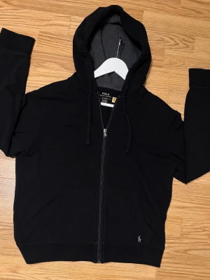 Svart hoodie från Polo Ralph Lauren - Snygg svart hoodie från Polo Ralph Lauren med dragkedja framtill, justerbar huva och fickor på magen. Klassisk logga broderad nedtill. Tillverkad i mjuk bomullsblandning som känns skön mot huden. Perfekt för en avslappnad och stilren look. Vid minsta fråga hör av dig mvh✌️