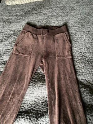 Bruna mjukisbyxor från Juciy Couture  - Mysiga bruna mjukisbyxor från Juciy Couture med bred resår i midjan och dragsko. Byxorna har två stora fickor fram och två bakfickor med knapp. Liten broderad logga på ena fickan framtill. Perfekta för en avslappnad stil. Ena snör saknar guld grejen, samt att sömnen i vänstra bakfickan gått upp.
