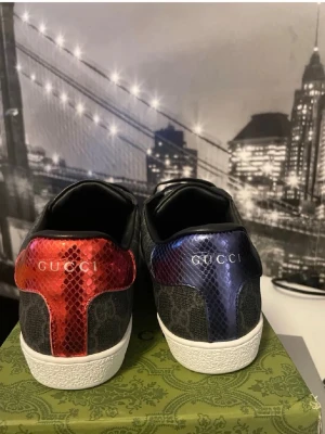 Gucci sneakers med GG-mönster - Snygga Gucci sneakers i svart med klassiskt GG-mönster. Skorna har detaljer i röd och blå metallic ormskinnslook på hälen, vita sulor och svarta skosnören. Märkeslogga på både häl och innersula. Perfekta för dig som vill sticka ut med exklusiv stil.